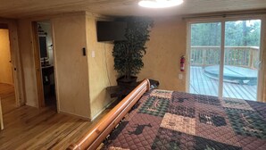 Ein Schlafzimmer mit Holzbalkendecke, einem großen Bett mit Steppdecke, einem an der Wand montierten Fernseher, einer Topfpflanze und einer Schiebeglasdoor zum Balkon.