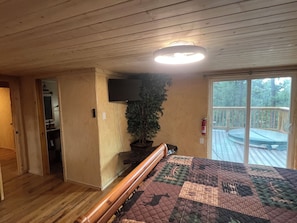 Un dormitorio con techo de madera, una cama grande con colcha, un televisor en la pared, una planta en maceta y un ventanal corredero que da a una terraza.