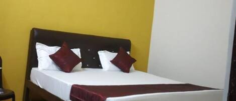 A single bed with a wooden headboard, white bedding, and two red pillows in a room with yellow walls and a white ceiling.