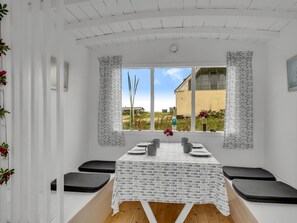 Un coin repas avec une table dressée pour quatre personnes, deux chaises de chaque côté, une fenêtre donnant sur un bâtiment et un paysage, et une nappe à motifs.