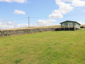 A single-story house with a green exterior and a wooden deck, situated on a grassy hill with a stone wall in the background. - The Lodge at Piece Farm (High Peak)
