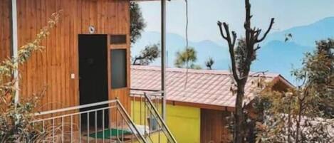 Una cabaña de madera con una escalera metálica que lleva a una puerta, rodeada de vegetación seca y un cielo despejado.