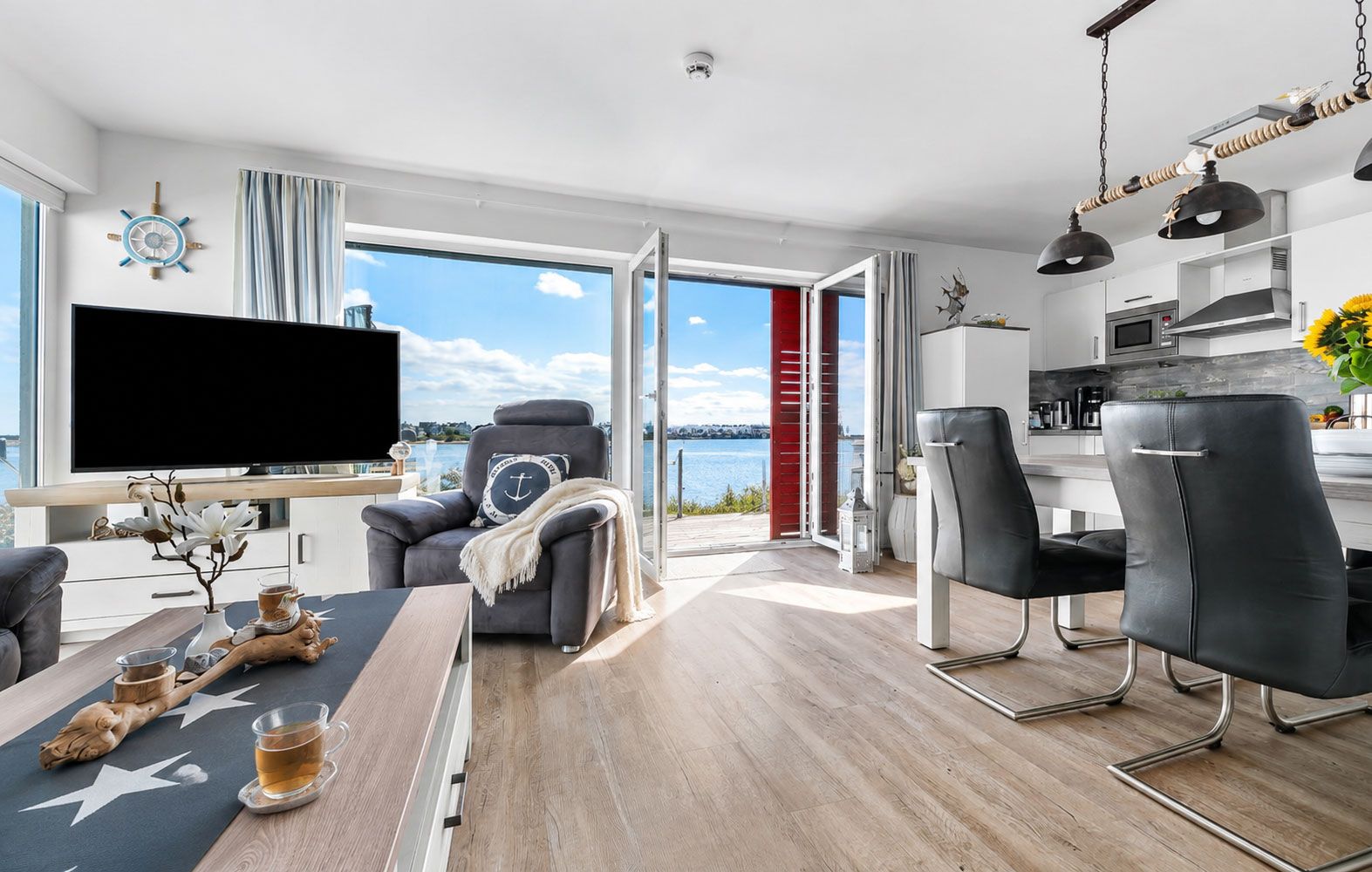 Uma sala de estar moderna com uma grande televisão de tela plana, uma área de estar confortável com sofá e poltronas, uma mesa de jantar com uma mesinha de centro e vista para o mar através de amplas janelas.