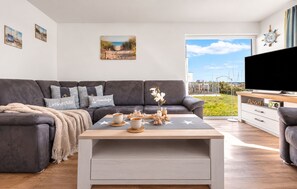 A modern living room with a grey sofa, a coffee table with decorative items, and a television on a stand. - At the Marina – Vacation Rental with Ocean View, Sauna, and Hot Tub (Olpenitz)