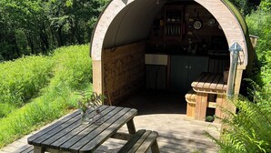 Eine hölzerne Außenküche mit einem Picknicktisch und Bänken, umgeben von viel Grün.