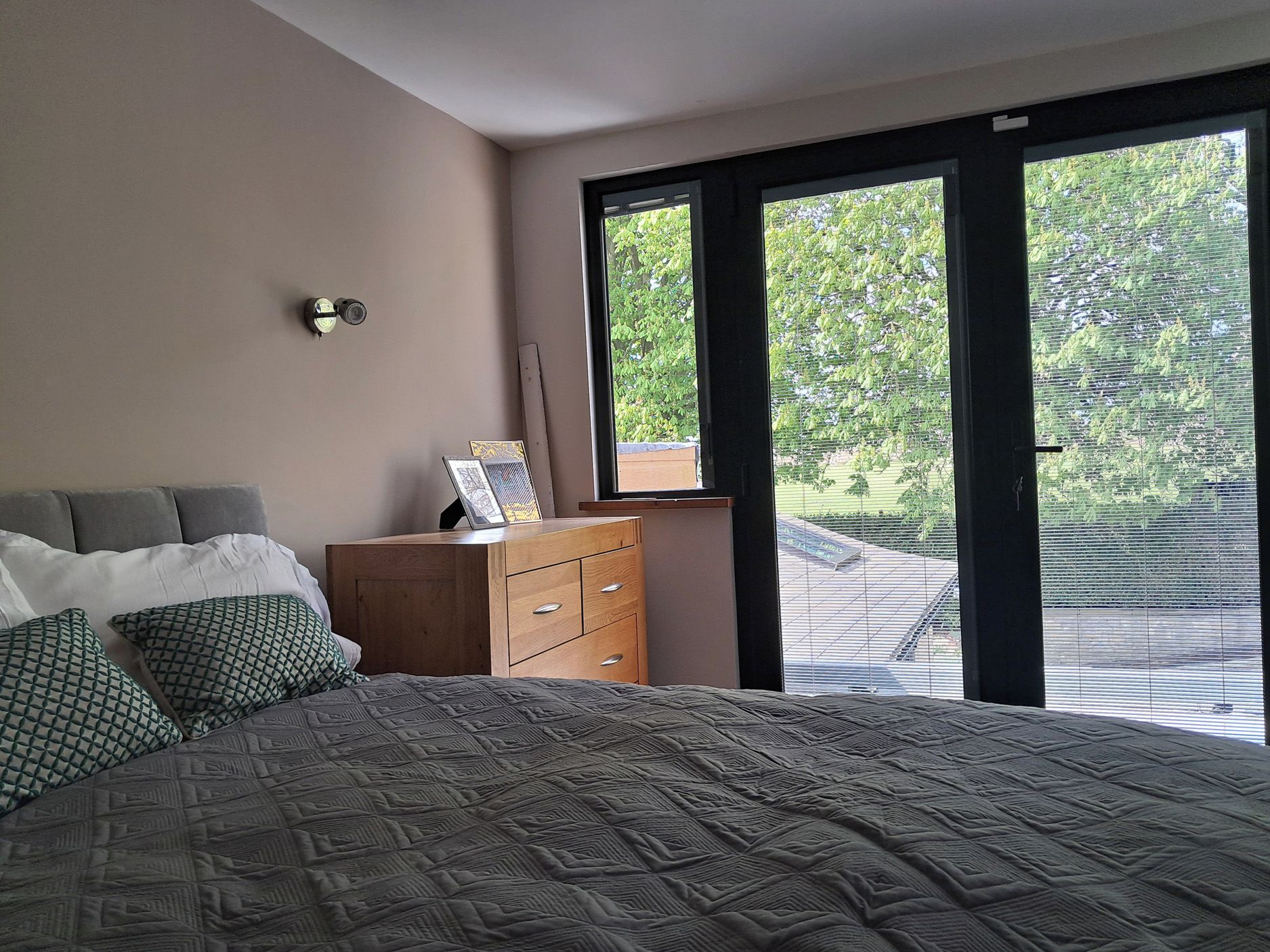 Een slaapkamer met een bed, een dressoir, een nachtkastje, een wandlamp en een raam met uitzicht op groen.