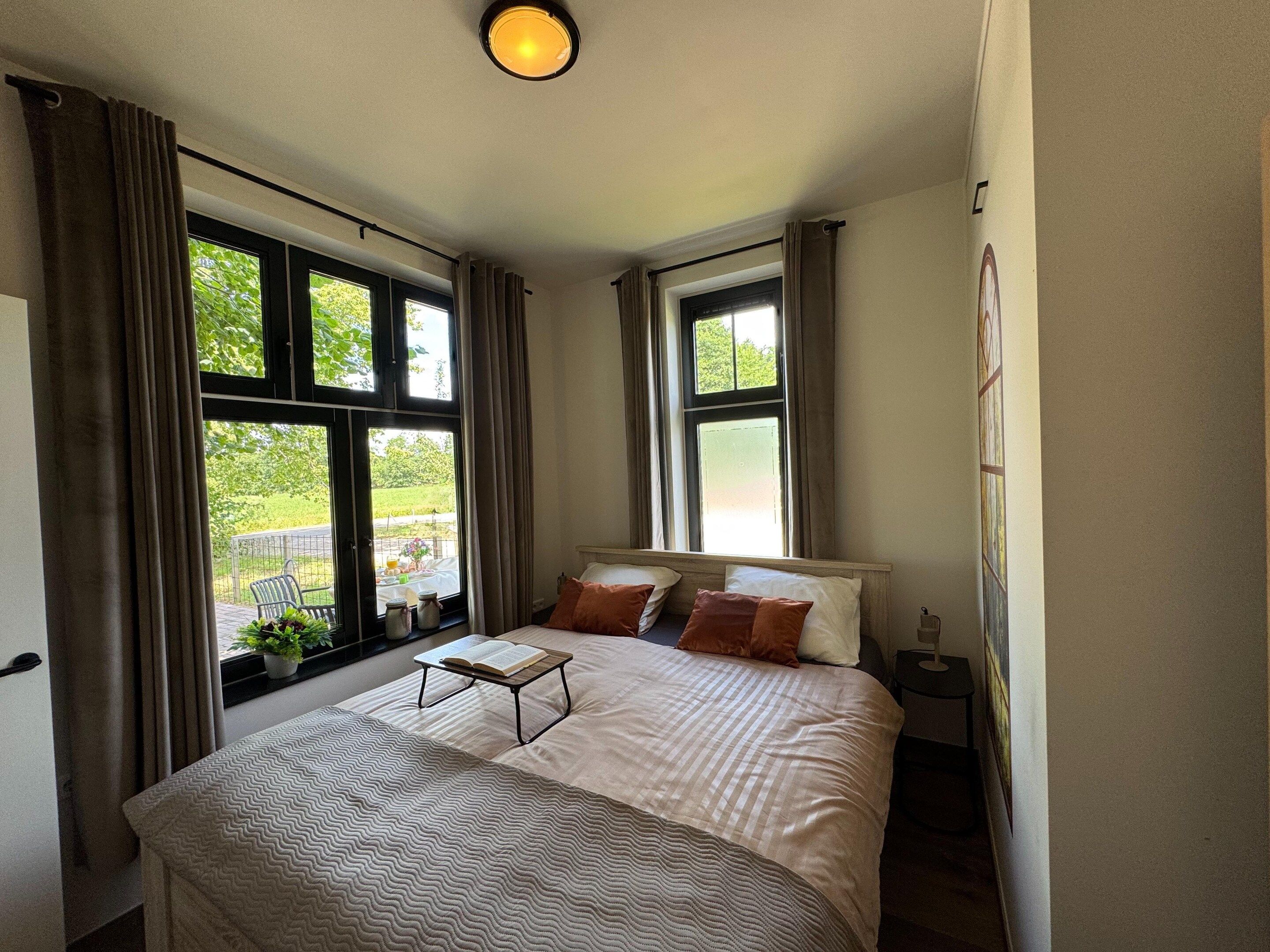 Un dormitorio con cama, mesita de noche, ventana con cortinas y vistas a zonas verdes.
