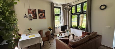 A dining area with a table, chairs, and a television. There is a sofa and a plant in the living area.
