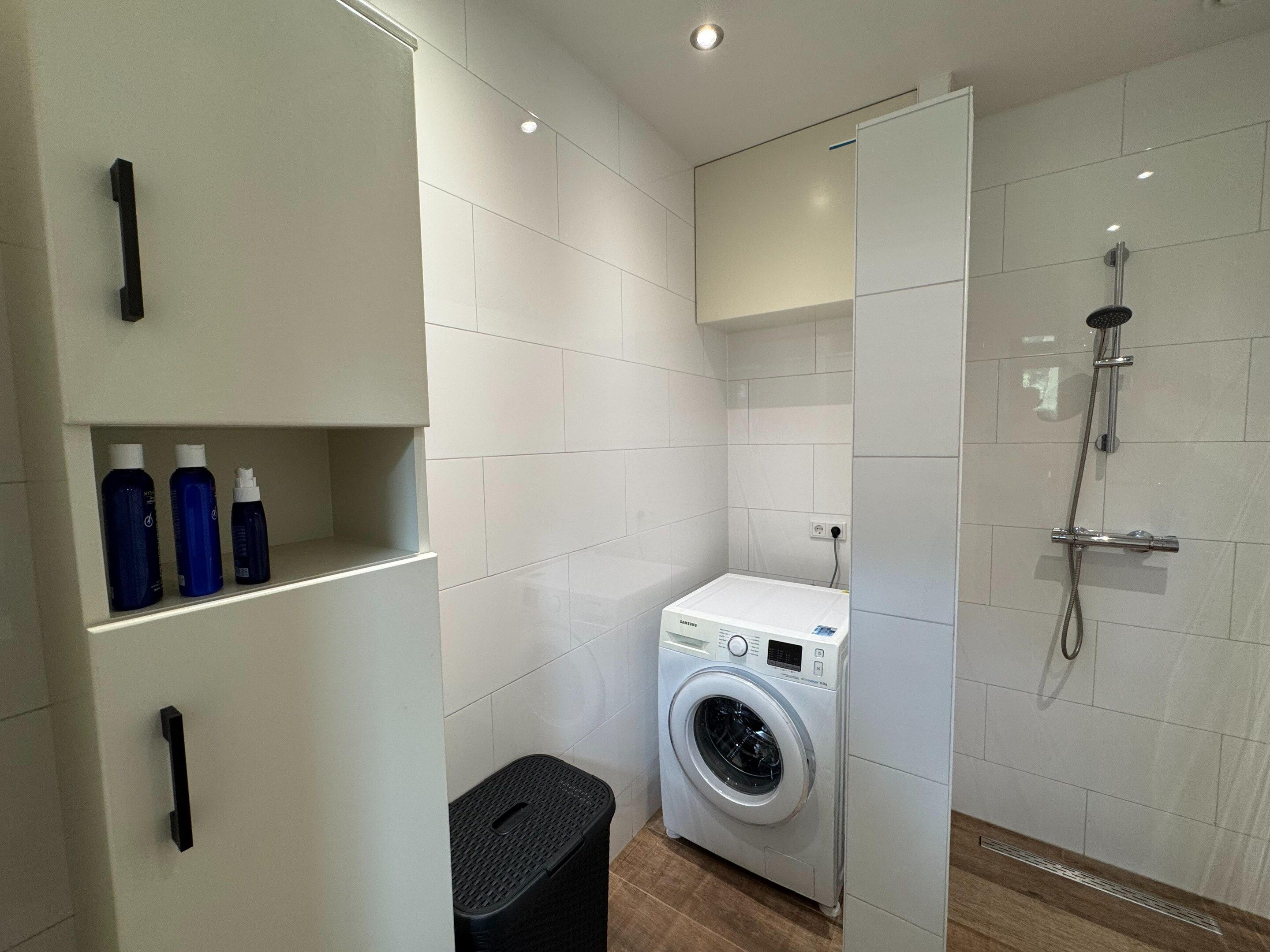 A compact bathroom with a washing machine, a shower, and a shelf with toiletries.