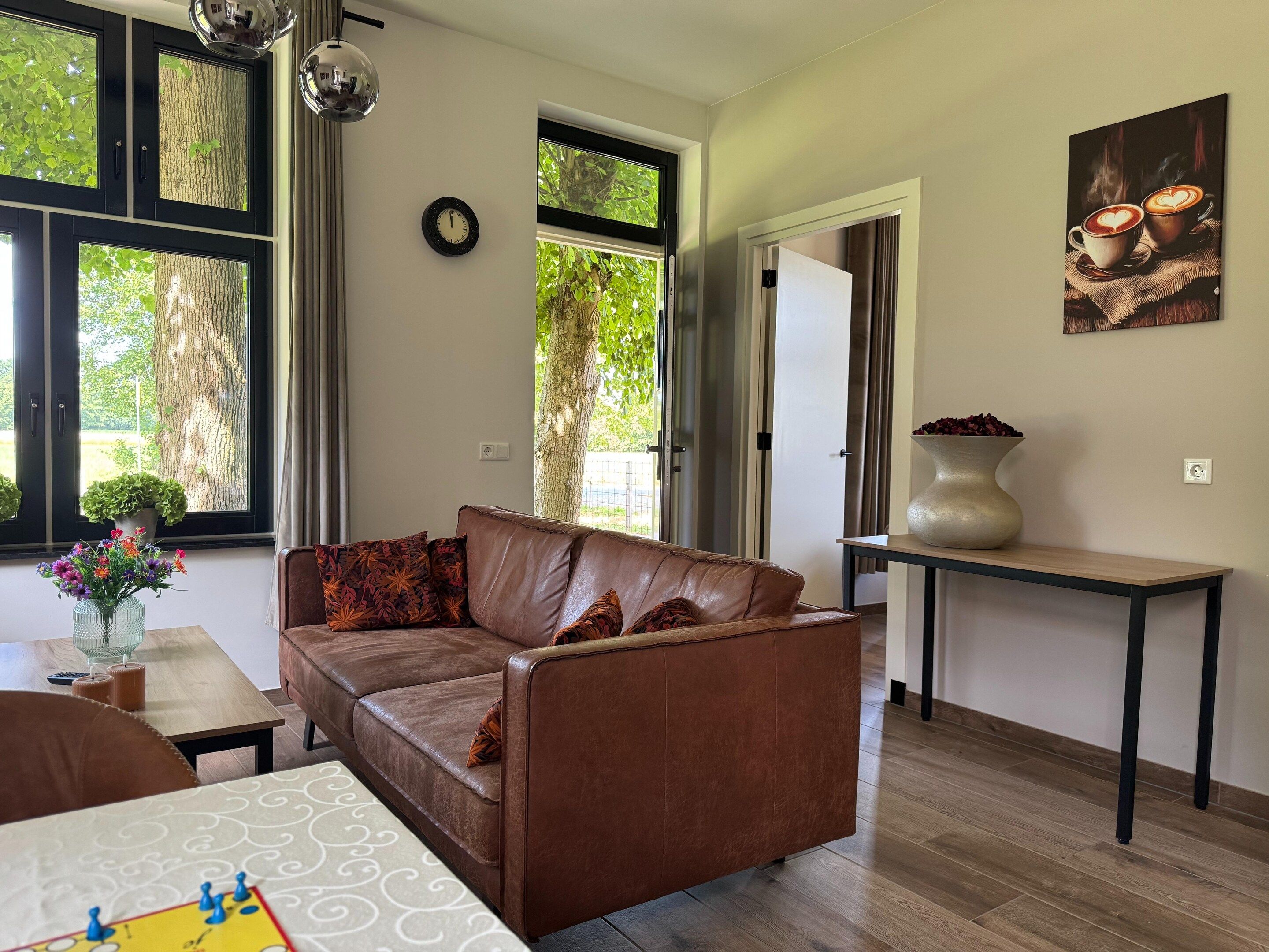 A modern living room with a brown leather sofa, a dining table with a chessboard, and a vase of flowers.