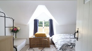 Ett sovrum med ett sluttande tak, en säng, en bänk, ett fönster med gardiner och ett litet bord med en vas med blommor.