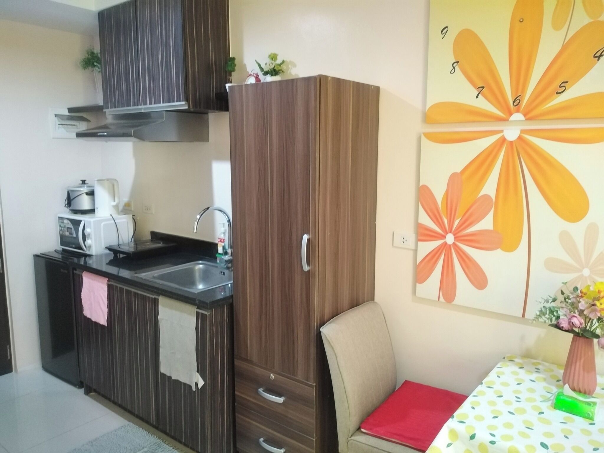 A compact kitchen with a microwave, toaster, and sink. There's a wooden cabinet with drawers and a small table with a red napkin and a green object on it.