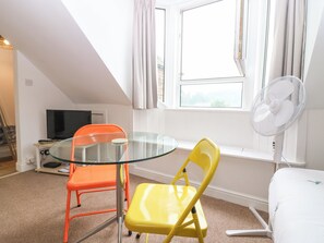 A room with a glass table, two chairs, a TV, a window with curtains, and a standing fan.