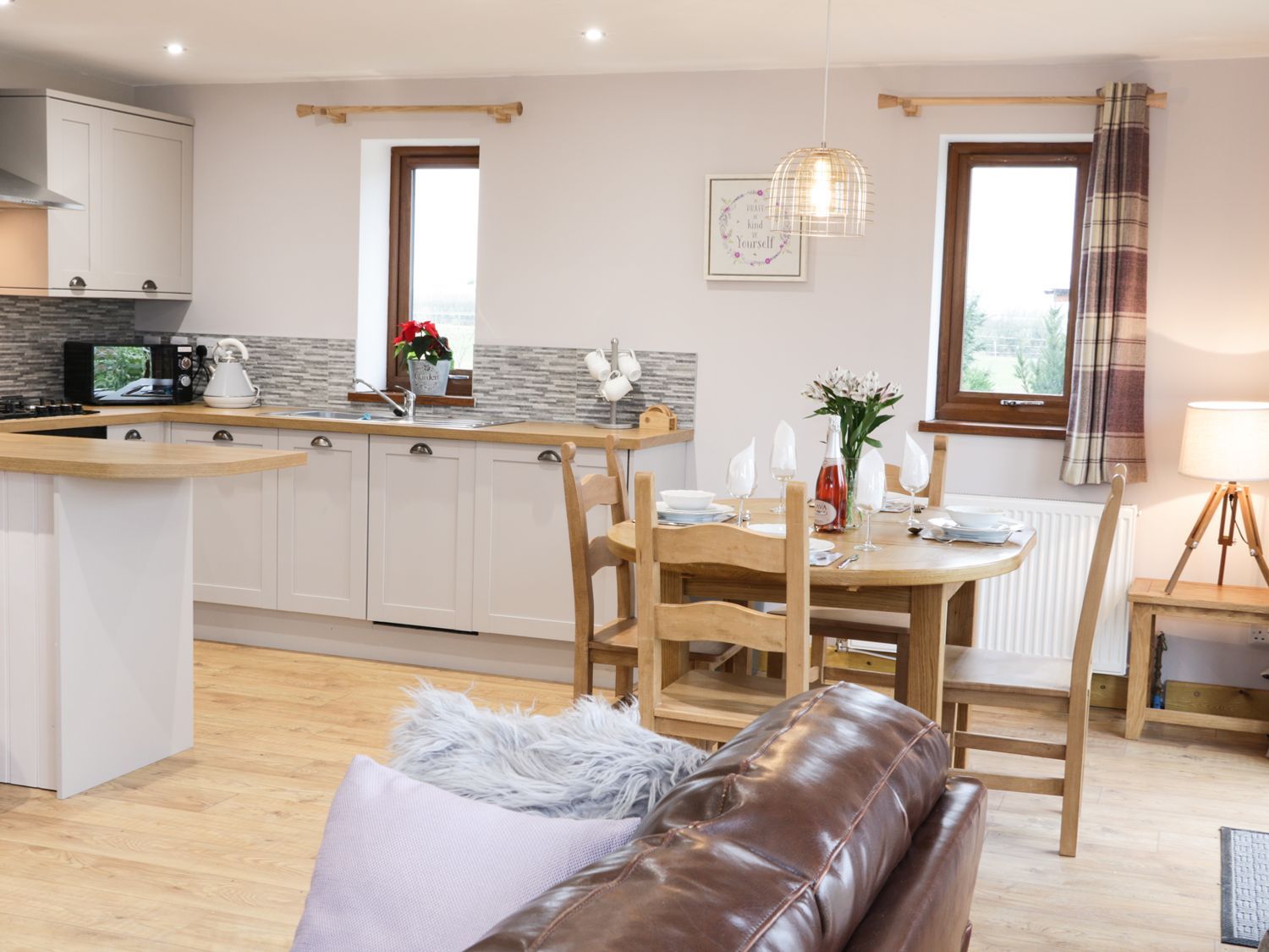 Een moderne keuken met witte kastjes, een ingebouwde magnetron en een waterkoker. Er is een eethoek met een houten tafel gedekt voor twee personen, een bruine leren bank en een zacht wit kussen.