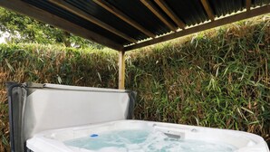 A hot tub under a covered area with a metal frame, surrounded by dense greenery.