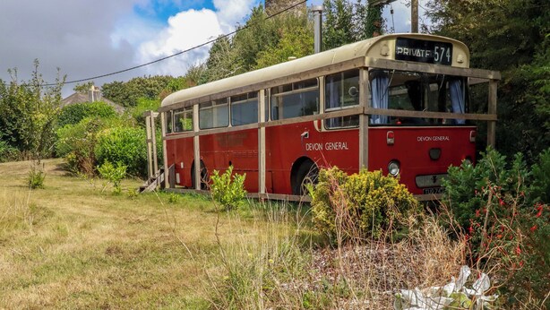 A red bus with 'DEVON GENERAL' written on the side parked in a grassy area with vegetation around.