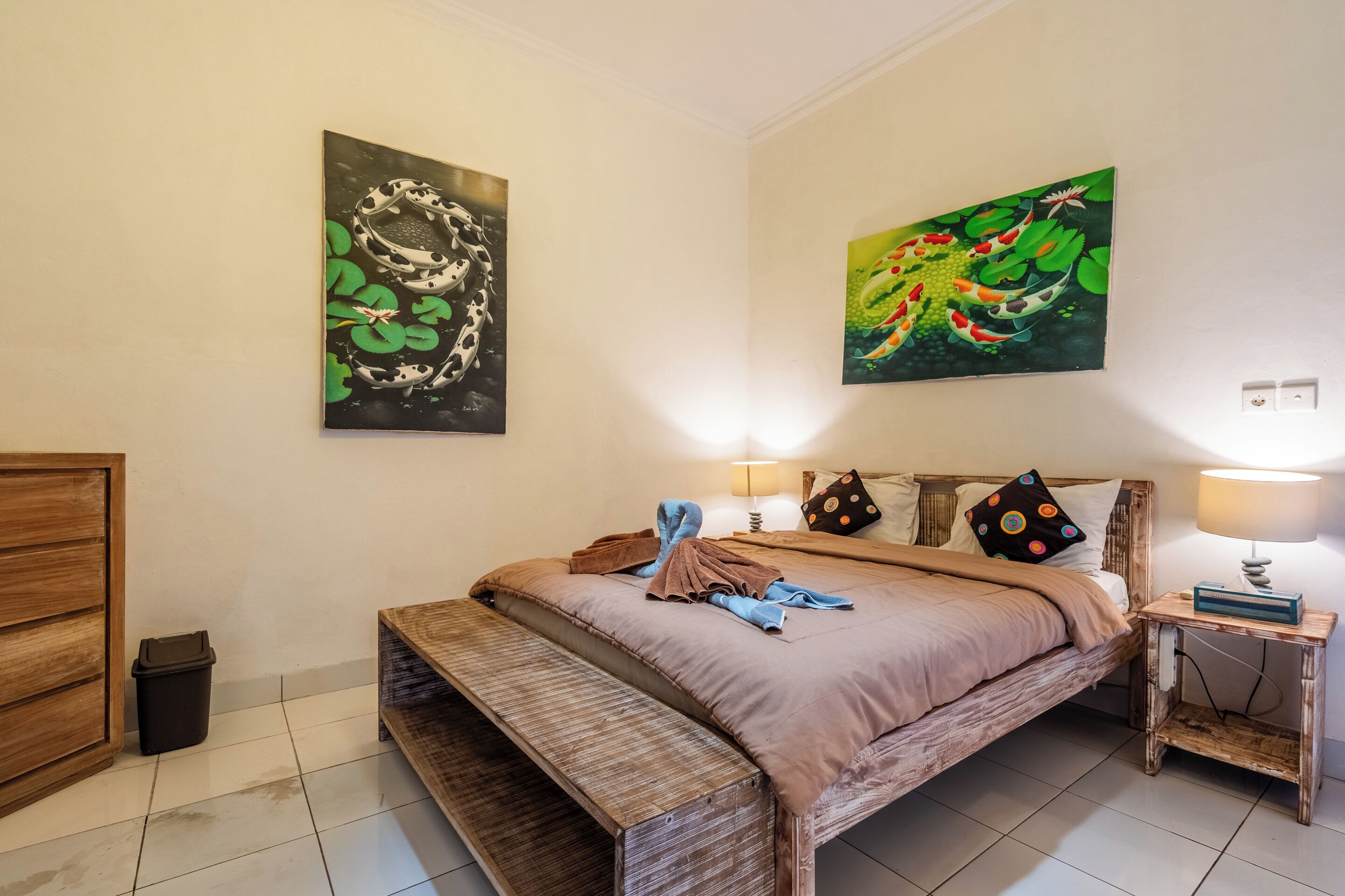 A bedroom with a bed, wooden bench, paintings, pillows, and a bedside table with a lamp.