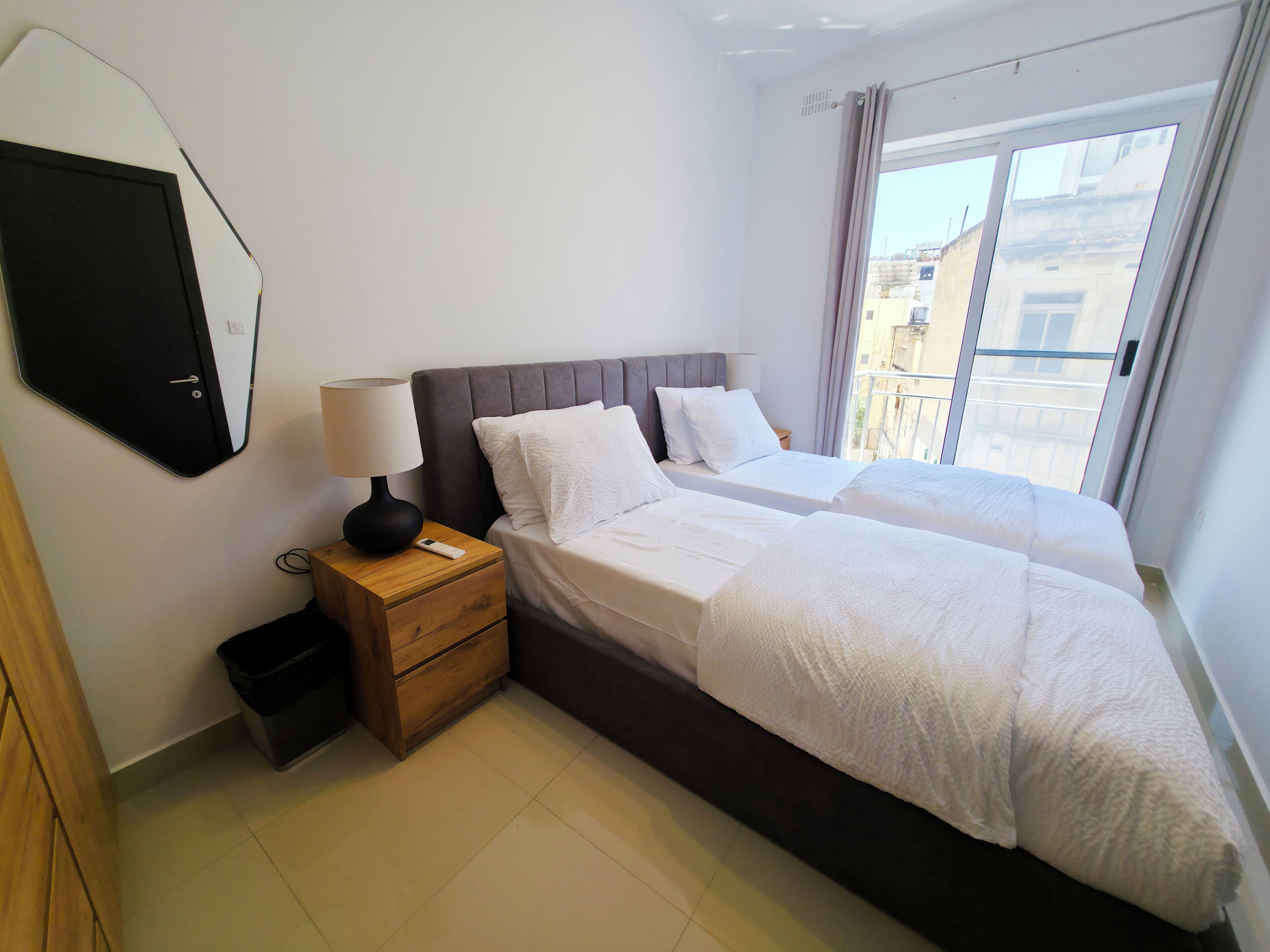 A bedroom with two beds, a nightstand with a lamp, a mirror, and a view of the outside.