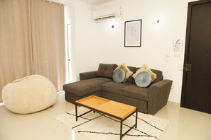 Una sala de estar con un sofá marrón, dos almohadas, una mesa de centro de madera, un pouf color beige y una alfombra blanca.