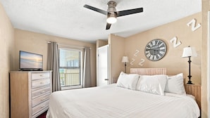 A hotel room with a bed, a television, a dresser, a ceiling fan, and a wall clock. - Beachfront condo w oversized balcony, pool & direct beach access (Gulf Shores)