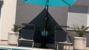 Un área de piscina con una sombrilla turquesa, dos sillones de descanso y plantas en macetas.