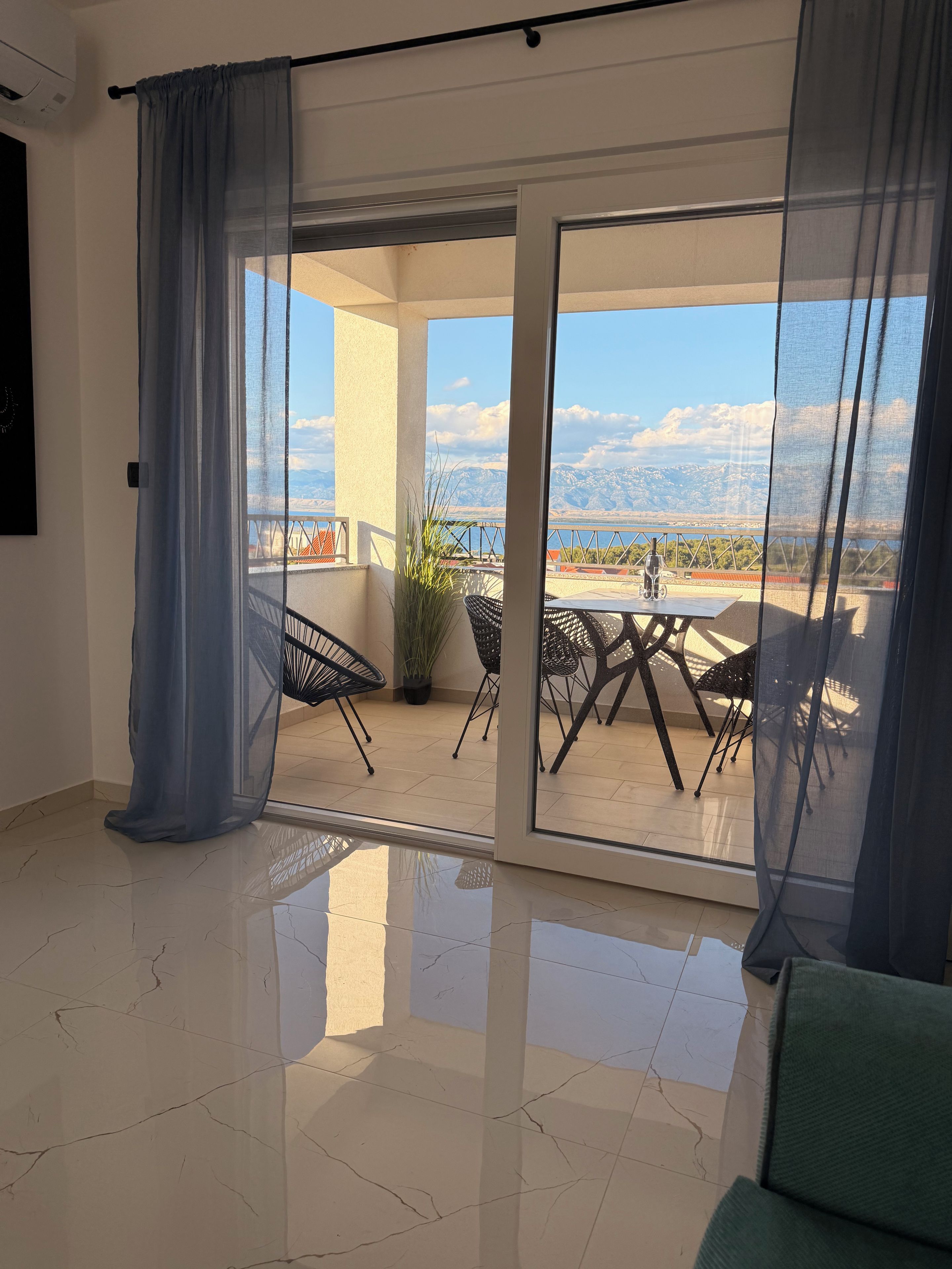 A room with a view of mountains and a bridge, a balcony with chairs and a table, and a sliding glass door.