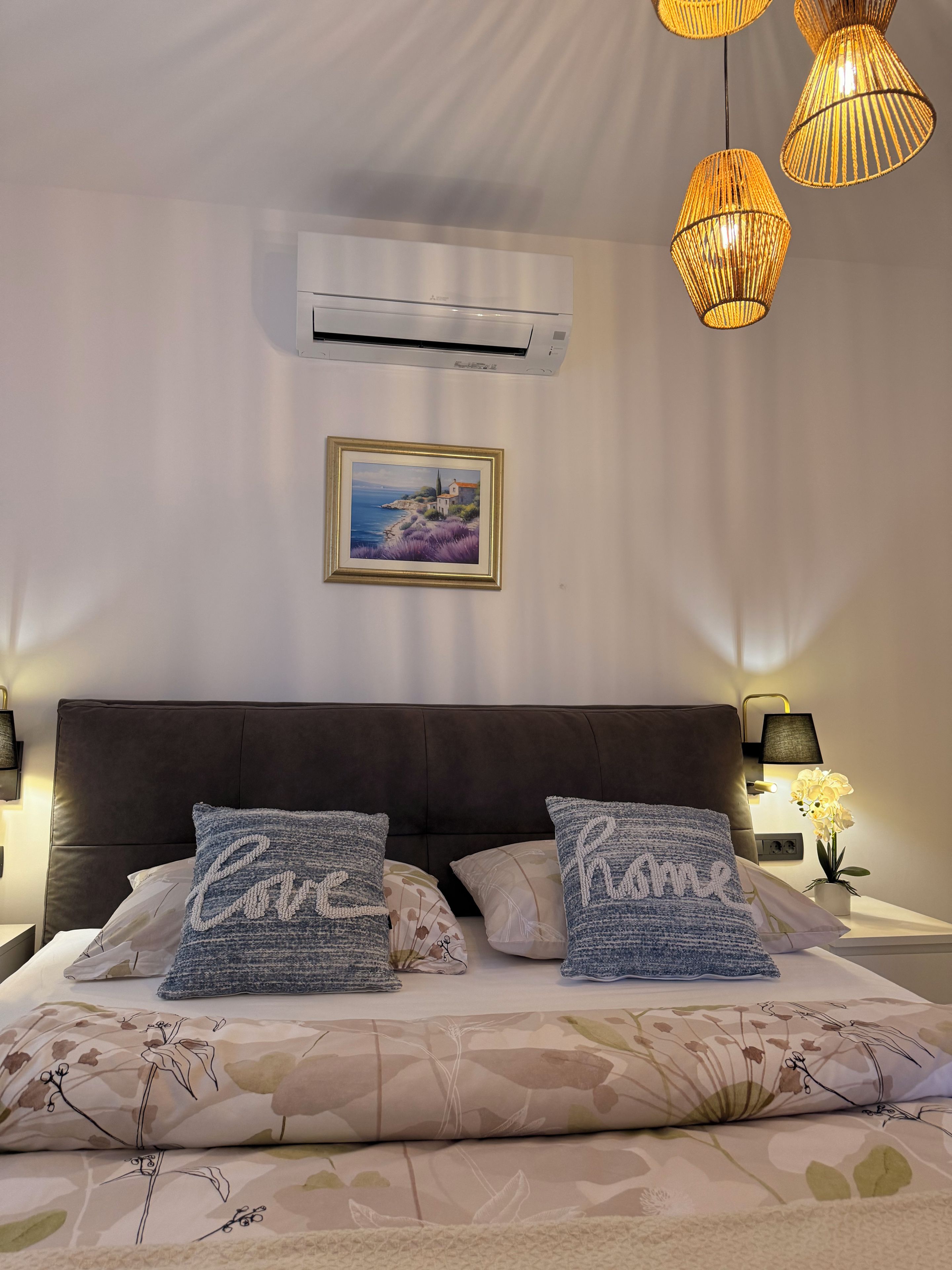 A bedroom with a large bed, two decorative pillows, a framed picture on the wall, an air conditioning unit, and three hanging lamps.
