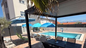 A poolside area with a table and chairs, umbrellas, and a view of a mountain range. - Luxury Blue Sky (Vir)