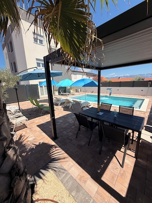 Un espace aménagé au bord de la piscine, avec une table et des chaises, des parasols et une vue sur une chaîne de montagnes.
