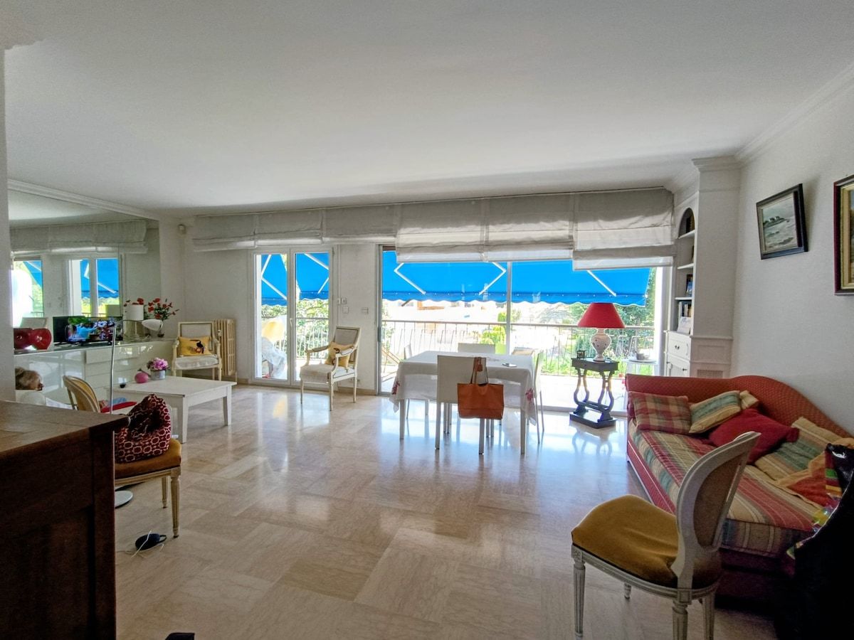 A spacious living area with a dining table, chairs, a sofa, and a view through large windows.