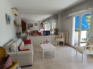 A spacious living area with a sofa, a coffee table, and a dining table. There is a kitchen area with a TV and various kitchen items. The room has large windows with a view of the outdoors. - Swimming pool, 3 bedrooms, baby-friendly (Toulon)