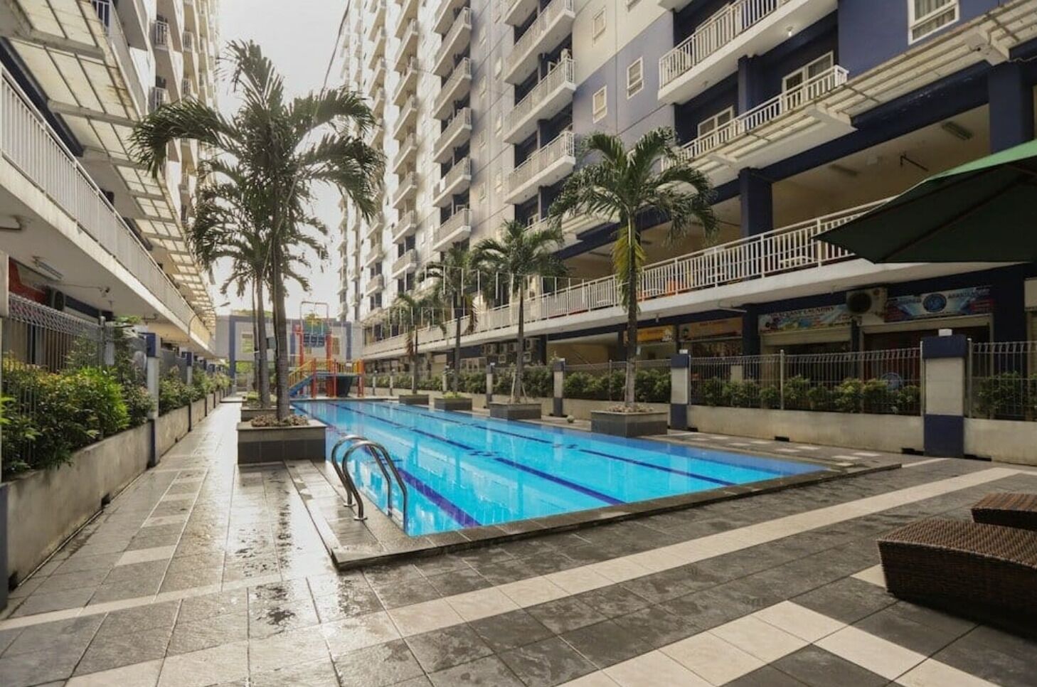 A swimming pool with a tiled deck, surrounded by multi-story buildings with balconies and palm trees.
