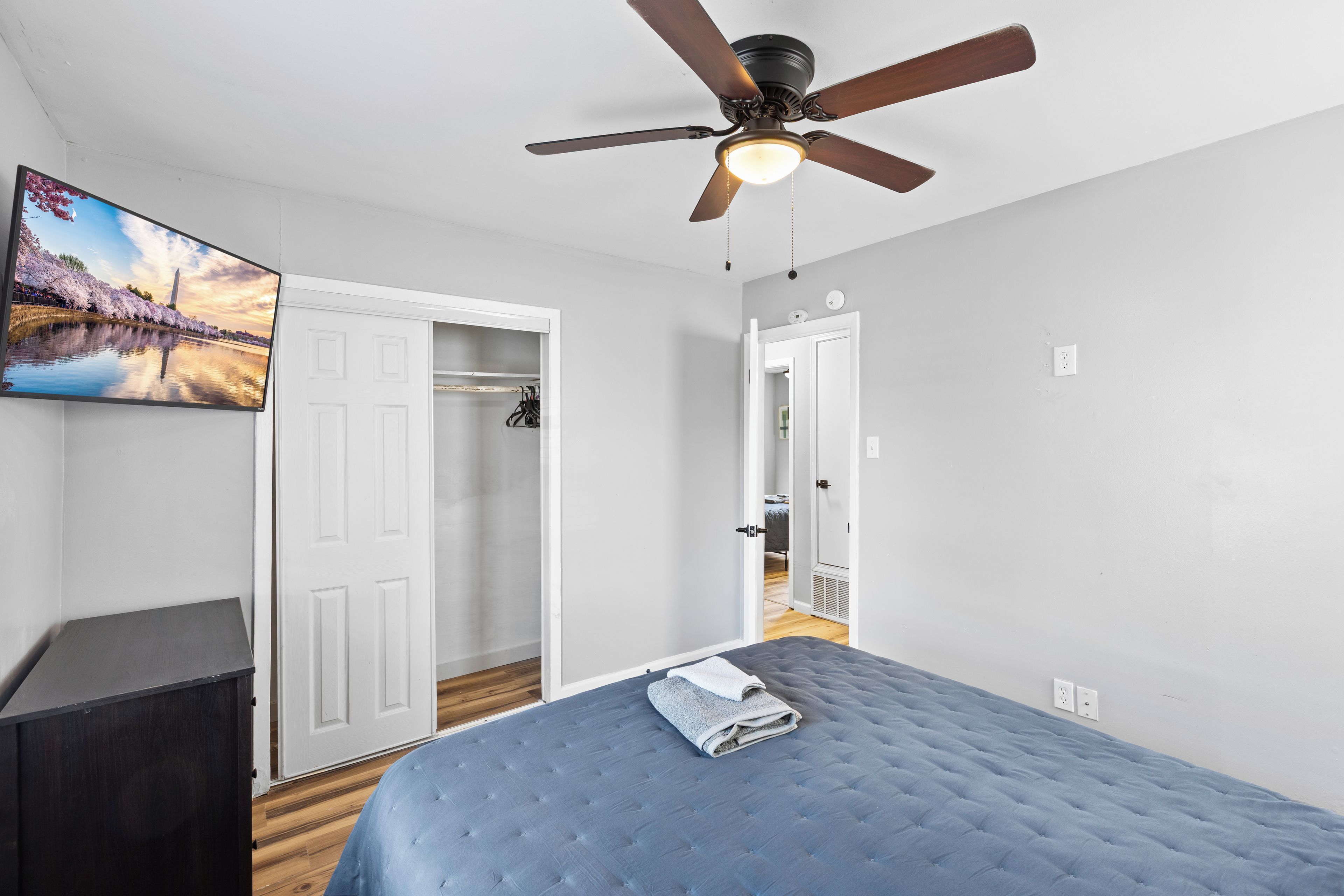 A bedroom with a bed, a ceiling fan, a door, and a wall-mounted TV.