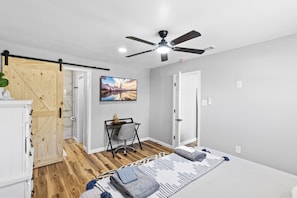 A bedroom with a bed, a desk with a chair, a ceiling fan, and a painting on the wall.
