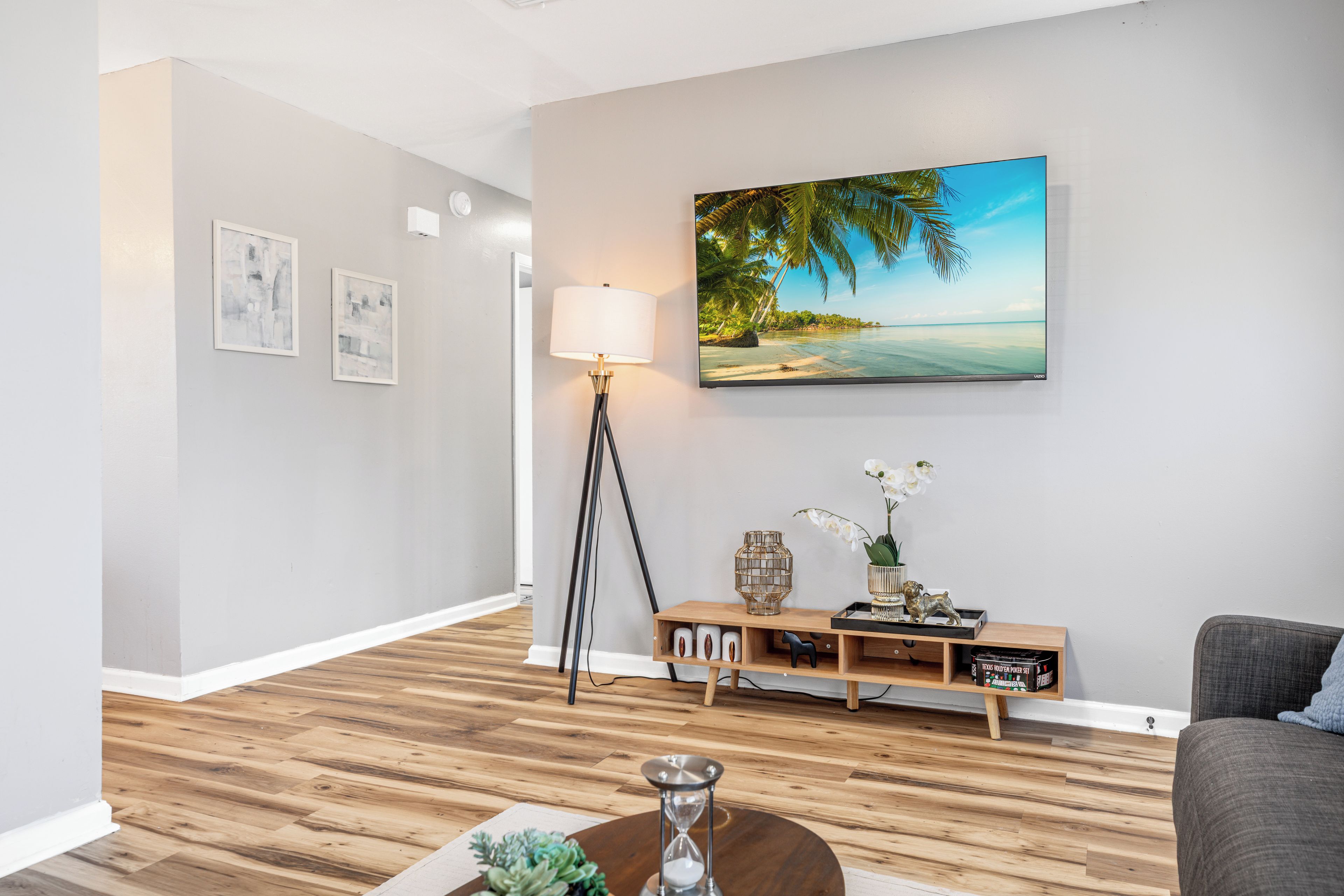 A modern living room with a wooden floor, a large painting of a beach, a floor lamp, and a console table with decorative items.