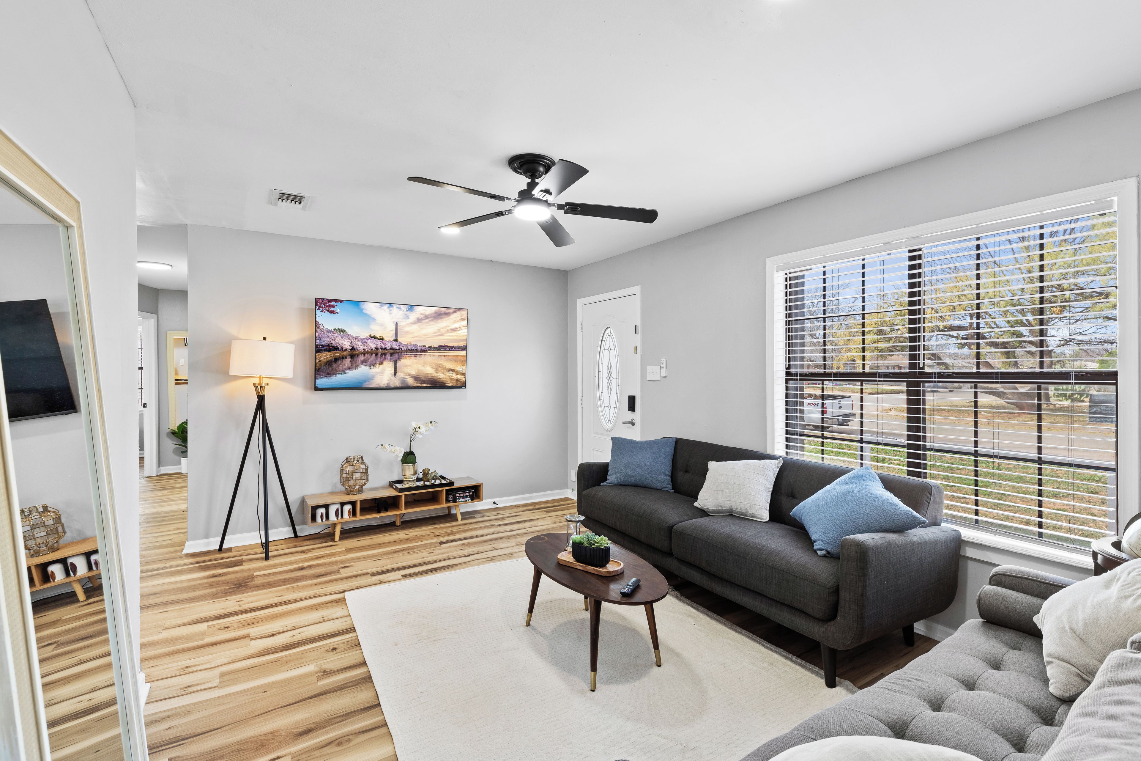 A modern living room with a dark grey sofa, a coffee table, and a large window with blinds.