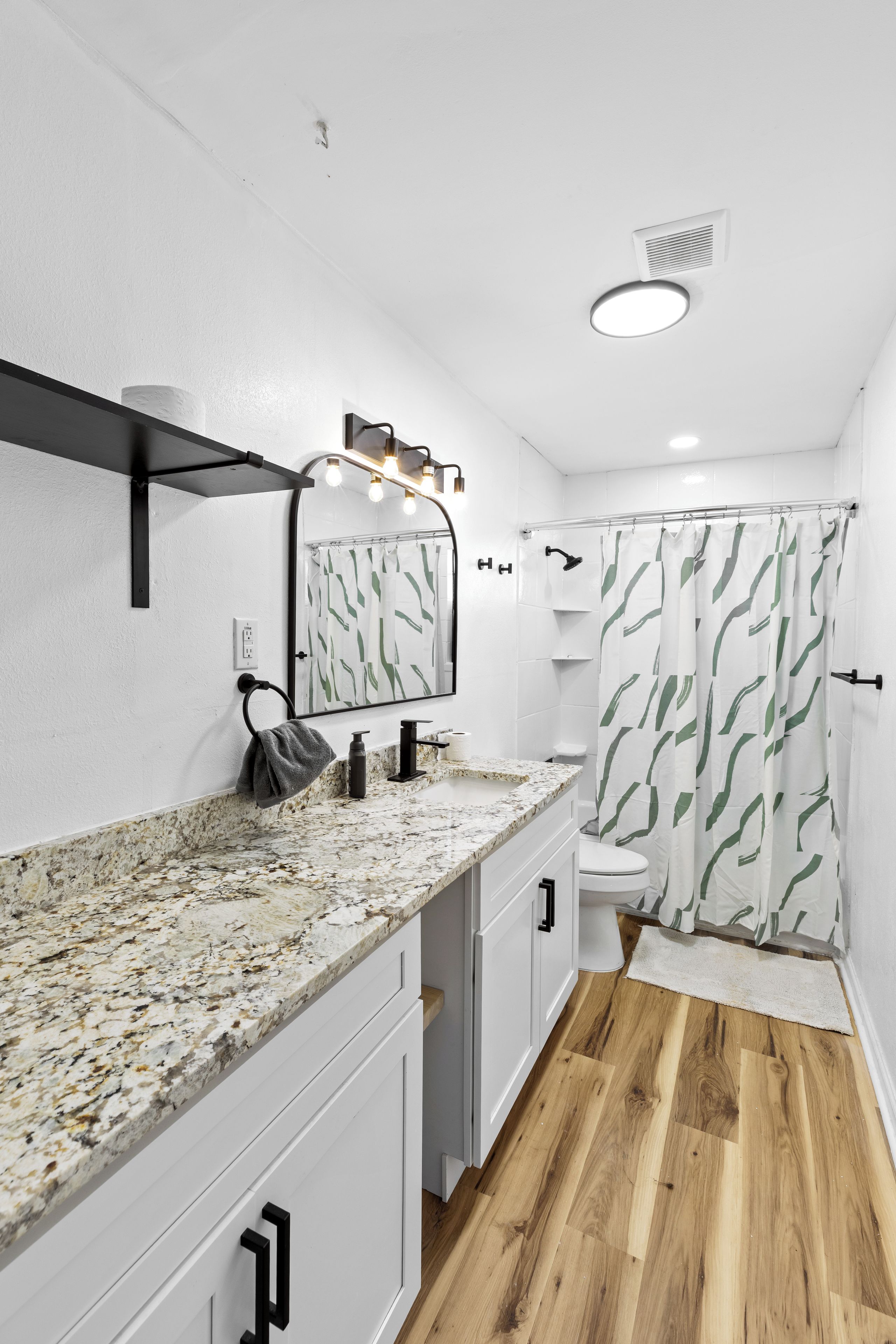 Un baño con encimera de mármol, un espejo con iluminación, una ducha con cortina de hojas verdes y suelo de madera.