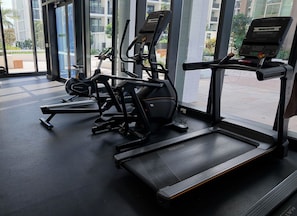 Ein Fitnessraum mit Cardiogeräten und Laufband, große Fenster mit Blick auf Gebäude und Grünflächen.