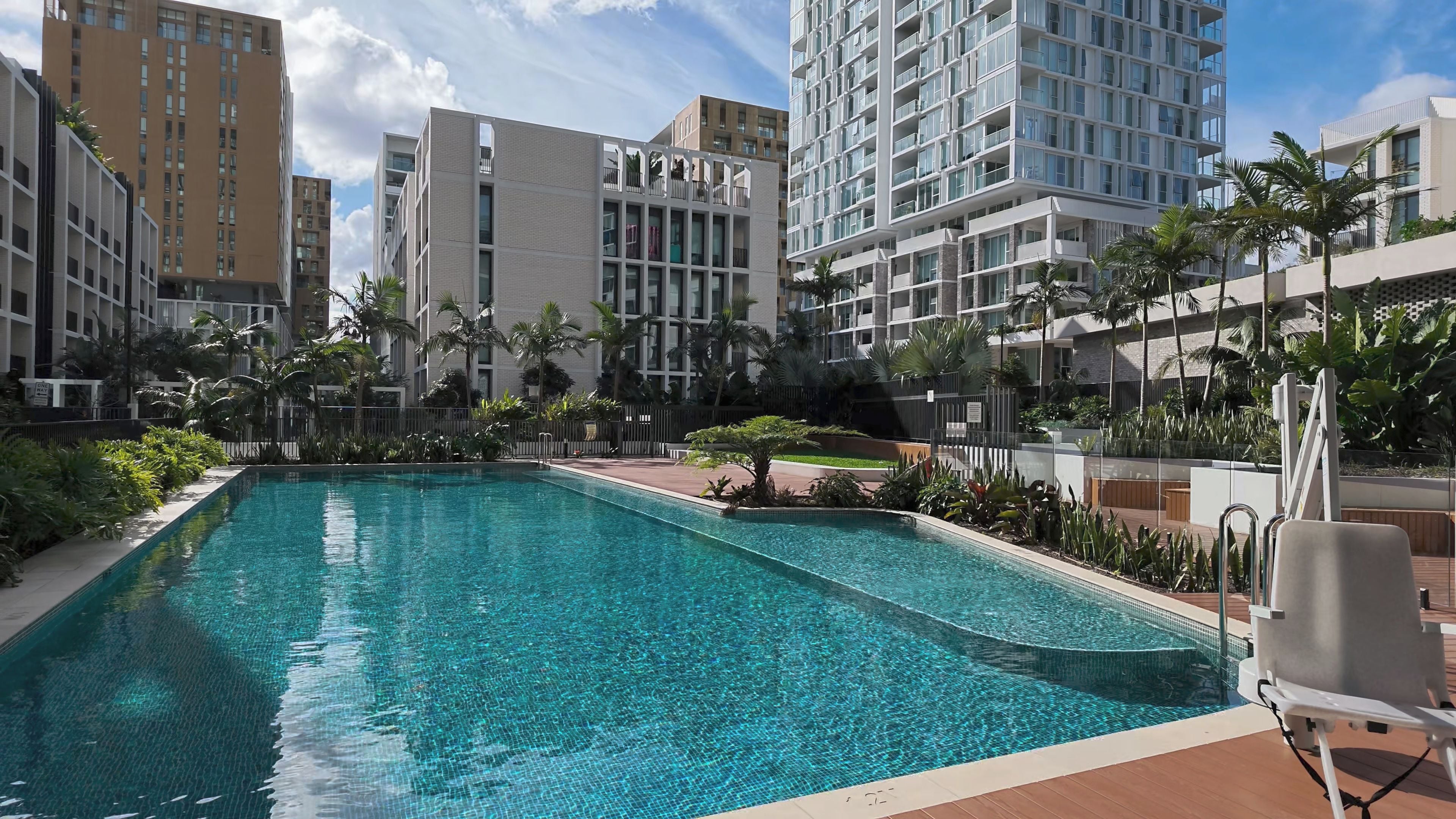 A swimming pool surrounded by modern buildings, palm trees, and a clear sky.