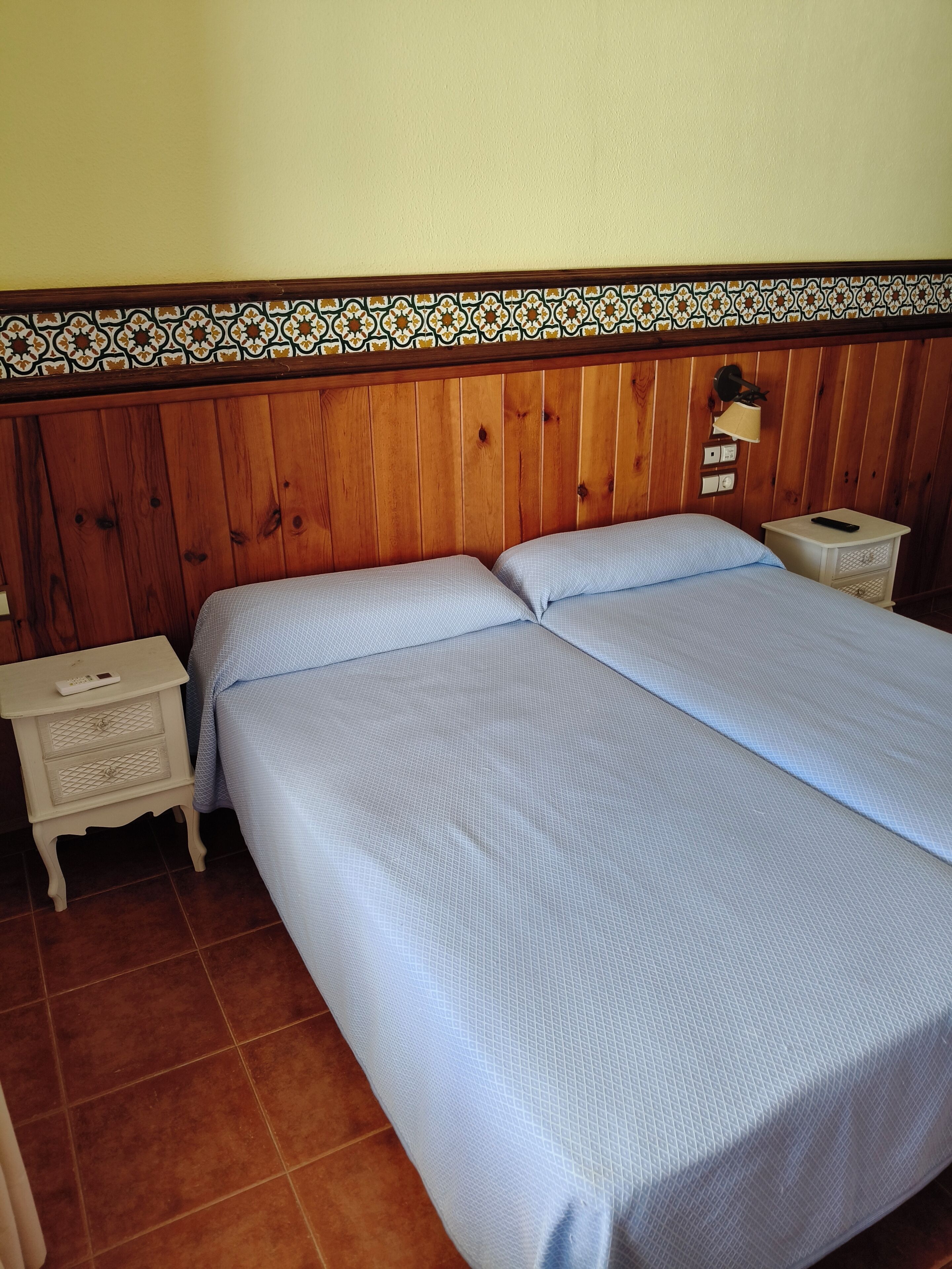 Une chambre avec une tête de lit en bois, deux lits avec une literie bleue, une table de chevet avec une lampe et un luminaire fixé au mur.