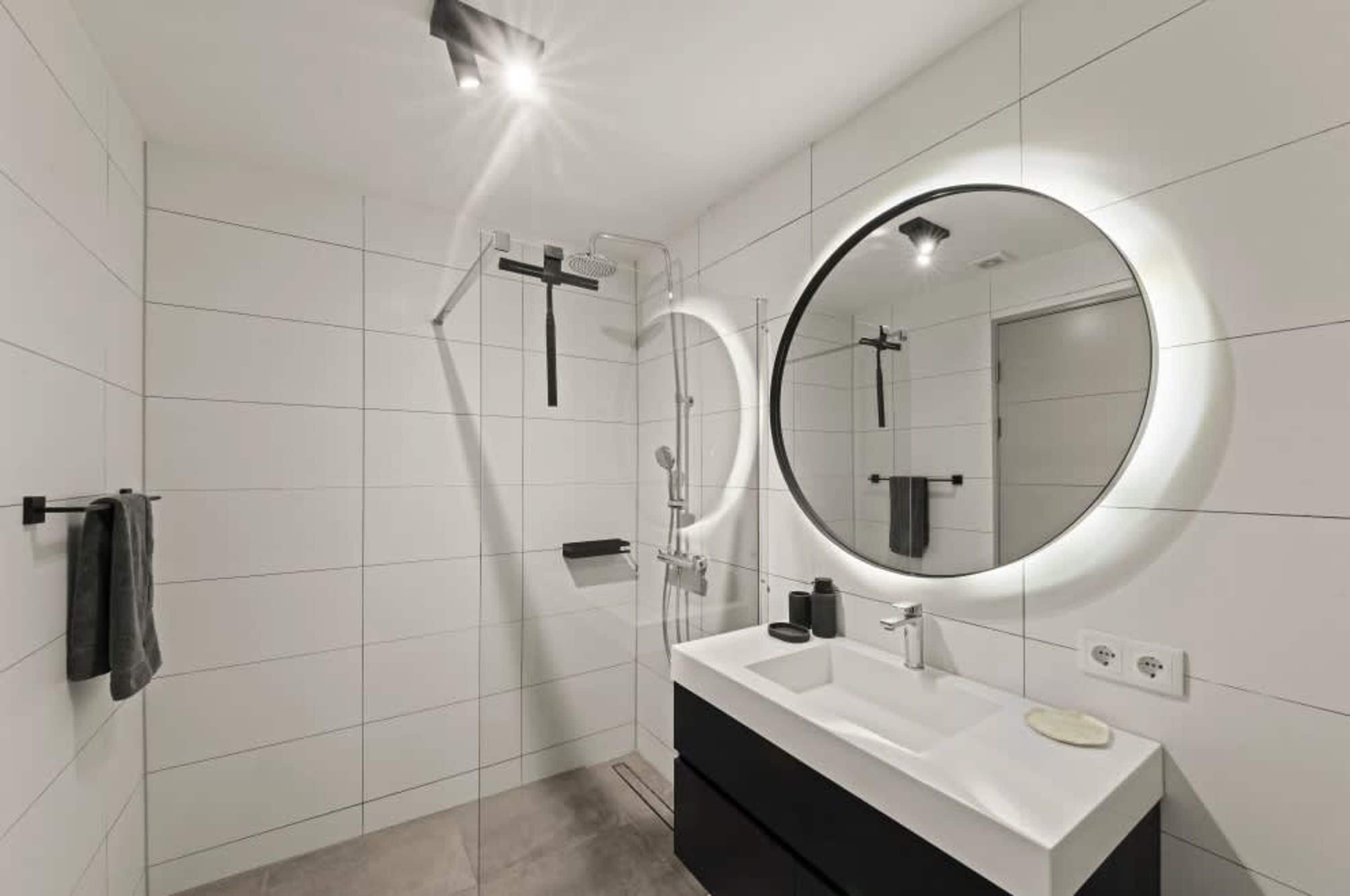 Un baño moderno con pared de azulejos blancos, un espejo redondo con luz, un tocador negro y una zona de ducha.