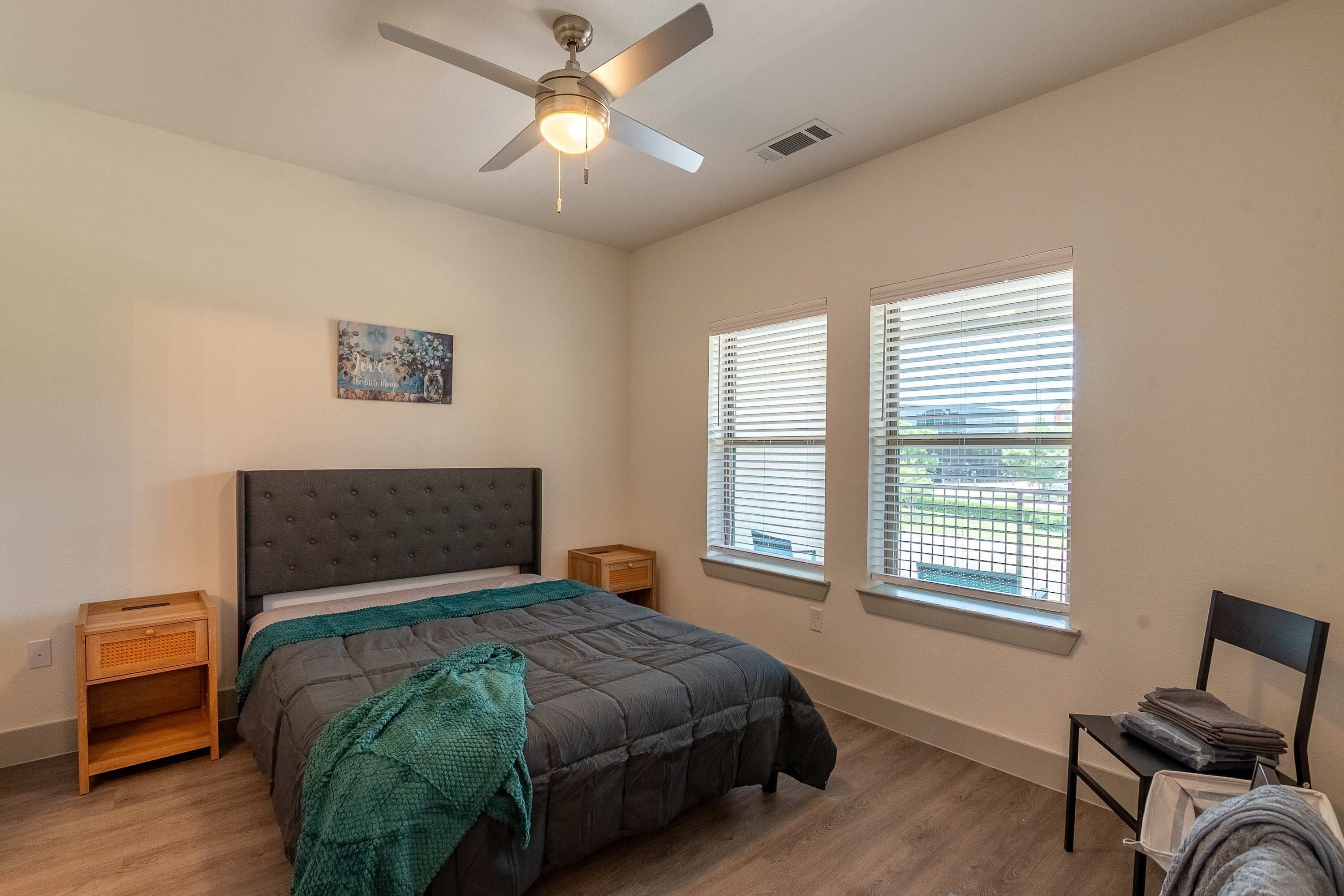 A bedroom with a bed, a ceiling fan, two bedside tables, a window with blinds, and a chair.