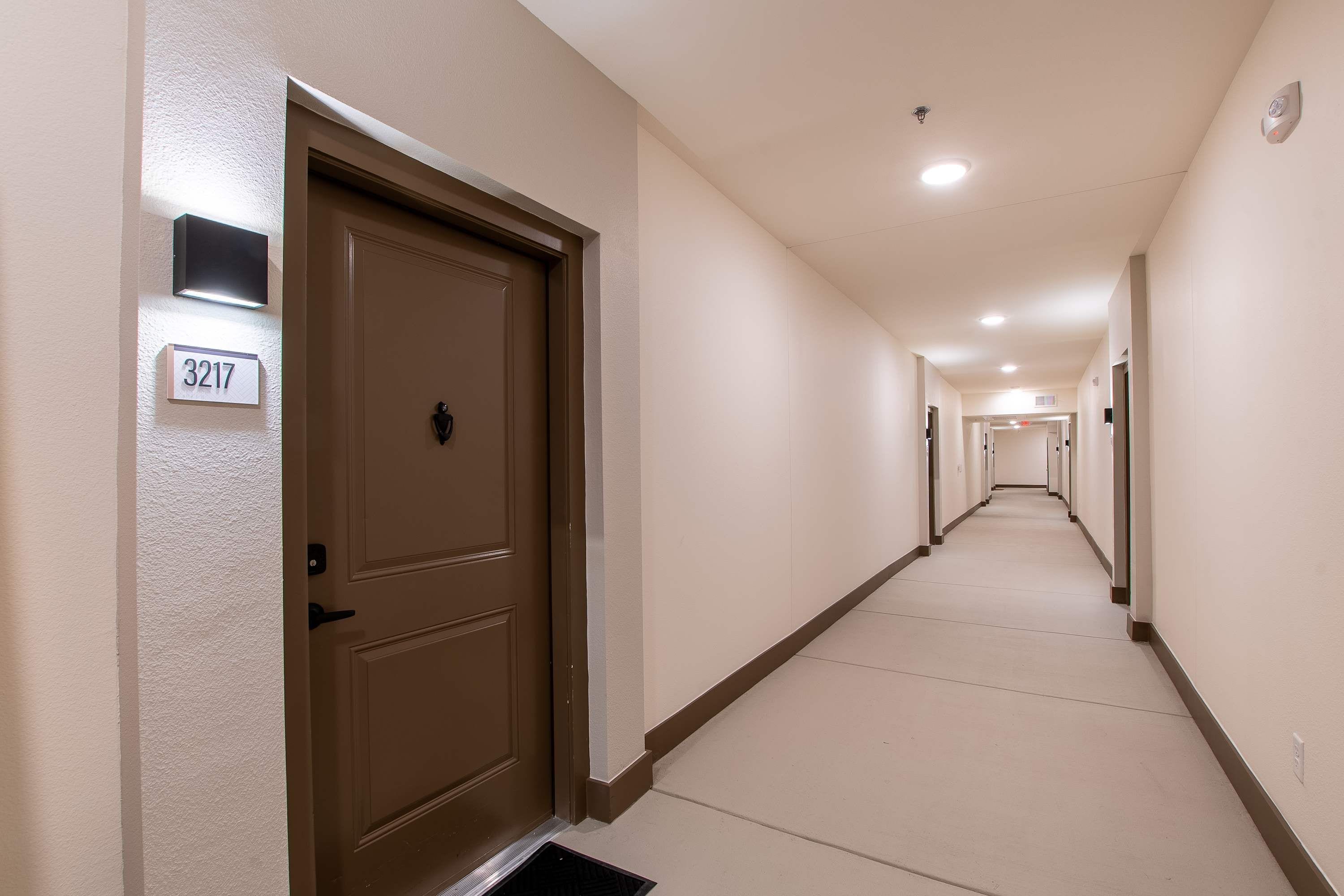 Un pasillo de hotel con una puerta marrón, un cartel con número y un largo pasillo que conduce a una puerta al final.