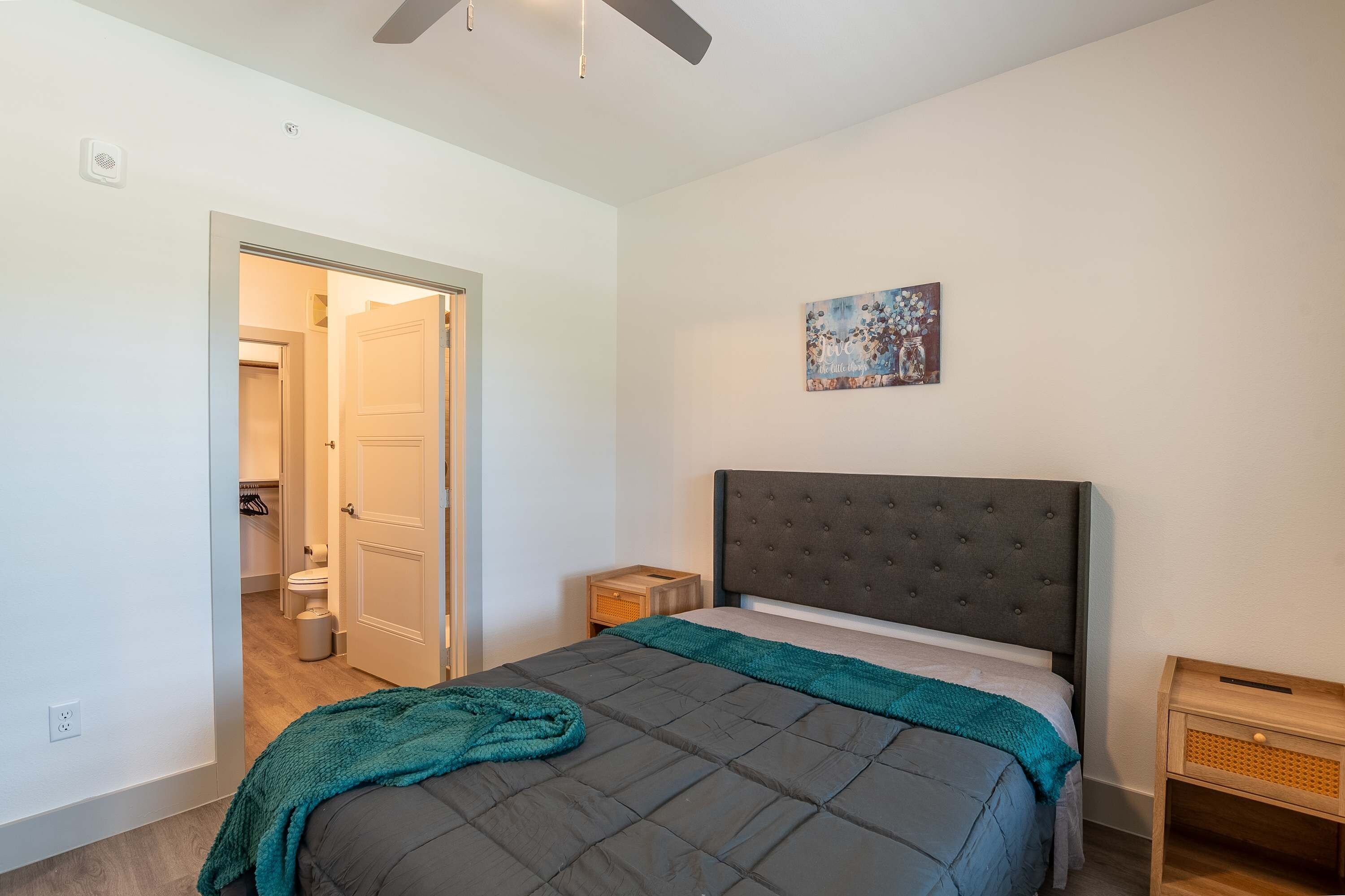 Un dormitorio con cama, mesita de noche, un cuadro en la pared y una puerta que da al baño.