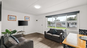 Uma sala de estar moderna com um sofá cinzento, uma televisão e uma janela com vista para uma área residencial.