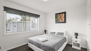 A bedroom with a large bed, two bedside tables, a window with blinds, and a framed picture on the wall. - Phillip Island Cottage 2 (Cowes)
