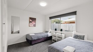 A bedroom with two beds, a desk, a window with a view of the outside, and a painting on the wall. - Phillip Island Cottage 2 (Cowes)