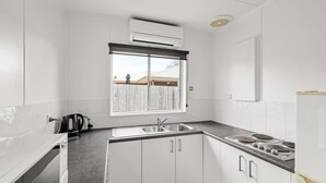 A kitchen with white cabinets, a double sink, a stove, and a window with a view of a fence and a building. - Phillip Island Cottage 3 (Cowes)