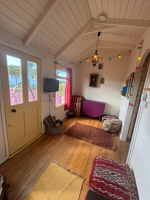Ein gemütliches Zimmer mit Holzbalkendecke, einem Fernseher, einem violetten Sofa und einem Fenster mit Blick auf ein blaues Haus und Palmen.
