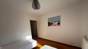 Une chambre avec deux lits simples, un plancher en bois, un tableau au mur et une porte donnant sur une autre pièce.